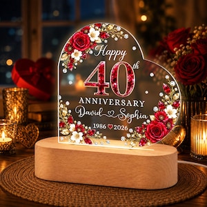 Könnte beinhalten: Eine herzförmige Acrylplatte auf einem hellen Holzsockel mit roten Rosen, weißen Blüten und goldenen Blättern. Der Text lautet: Happy 40th ANNIVERSARY David and Sophia 1986 and 2026. Die Zahl 40 ist groß, rot und hat eine edelsteinartige Textur.