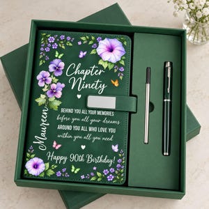 Könnte beinhalten: Ein dunkelgrünes Notizbuch-Set mit Magnetverschluss, passendem Stift und einem schlanken Eingabestift. Der Einband zeigt violette Blumen, Schmetterlinge sowie den Text Chapter Ninety, Maureen und Happy 90th Birthday mit Zitat.