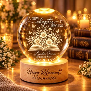 May include: A spherical glass night light on a round wooden base. The glass features an etched open book with flowers, butterflies, and the text "A NEW CHAPTER BEGINS", "Linda Jane", and "RETIRED 2027". The wooden base is engraved "Happy Retirement!".