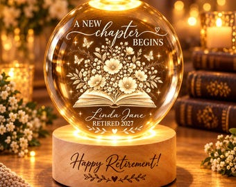 May include: A spherical glass night light on a round wooden base. The glass features an etched open book with flowers, butterflies, and the text "A NEW CHAPTER BEGINS", "Linda Jane", and "RETIRED 2027". The wooden base is engraved "Happy Retirement!".