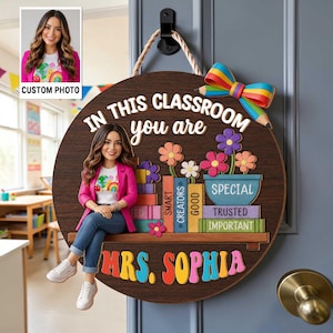 May include: A round wooden door sign featuring a cartoon illustration of a woman in a pink blazer and blue jeans sitting on a shelf. The sign displays the text "IN THIS CLASSROOM you are" above colorful book spines and "MRS. SOPHIA" below.
