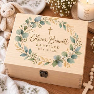 May include: A rectangular light-coloured wooden keepsake box featuring a metal latch and a printed design of a green eucalyptus wreath with gold accents. The text reads Oliver Bennett BAPTIZED MAY 17, 2026, centred below a small gold cross.