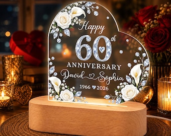 May include: A heart-shaped acrylic plaque on a light wood base featuring the text Happy 60th ANNIVERSARY David and Sophia 1966-2026. The design includes white roses, calla lilies, and sparkling diamond-like accents printed on the clear surface.