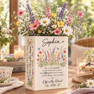 May include: A rectangular white ceramic vase shaped like a book, filled with colorful wildflowers. The front features the name Sophia, a floral illustration, and text reading: May you be proud of the work you've done, the difference you've made, and the lives you've touched. Officially Retired 2026. The side reads: A New Chapter Begins.