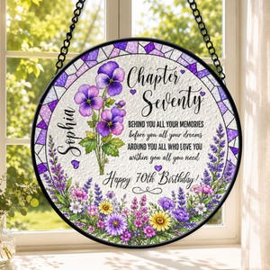 May include: A circular hanging sign with a stained-glass style purple border. It features purple pansies, lavender, and wildflowers. The text reads: Sophia, Chapter Seventy, BEHIND YOU ALL YOUR MEMORIES before you all your dreams AROUND YOU ALL WHO LOVE YOU within you all you need Happy 70th Birthday!