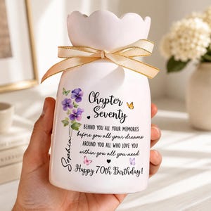 May include: A white ceramic vase with a scalloped rim and a beige ribbon tied around the neck. The vase features purple pansy flowers, butterflies, and the text: Chapter Seventy, BEHIND YOU ALL YOUR MEMORIES, before you all your dreams, AROUND YOU ALL WHO LOVE YOU, within you all you need, Sophia, Happy 70th Birthday!