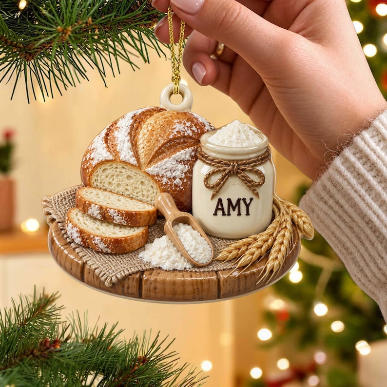 Op de afbeelding: Een kerstornament met een ronde houten plank, een gesneden brood, een klein potje met het label AMY gevuld met witte bloem, een houten schepje en tarwearen. Het ornament hangt aan een gouden koordje.