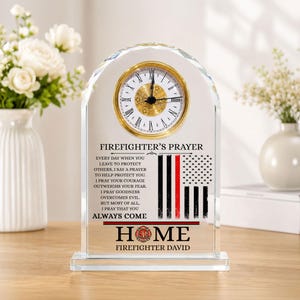 Puede incluir: Un reloj de sobremesa de cristal arqueado con esfera analógica de borde dorado y números romanos. La superficie de cristal muestra el texto FIREFIGHTER'S PRAYER, un gráfico de una bandera con una franja roja y la palabra HOME con un emblema.
