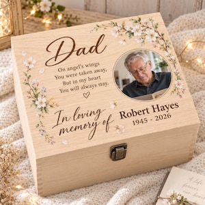 May include: A light-coloured wooden keepsake box featuring a circular photo of an older man, floral illustrations, and the text: Dad, On angel's wings You were taken away, But in my heart You will always stay, In loving memory of Robert Hayes 1945-2026.