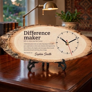 May include: A live edge wood slice clock featuring the printed text Difference maker, a definition, and the name Sophia Smith. The clock face has black hour and minute hands with a red second hand, supported by a black metal display stand.