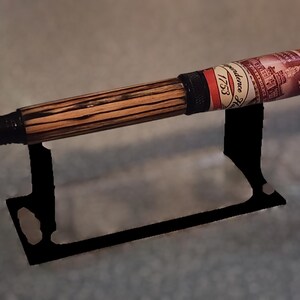 May include: A handcrafted pen with a wooden barrel featuring a striped pattern. The pen has a black tip and a decorative label with text and a red, white, and black design. It rests on a black metal stand.
