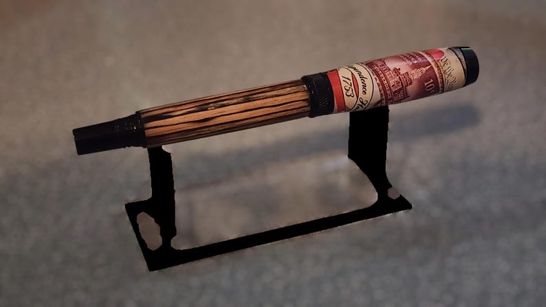 May include: A handcrafted pen with a wooden barrel featuring a striped pattern. The pen has a black tip and a decorative label with text and a red, white, and black design. It rests on a black metal stand.