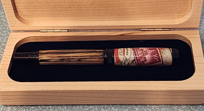 May include: A wooden pen with a striped pattern and a stamp-themed design, presented in a light-colored wooden box with a black velvet interior. The pen features the text "Providence Hall 1753" and "10&cent;".