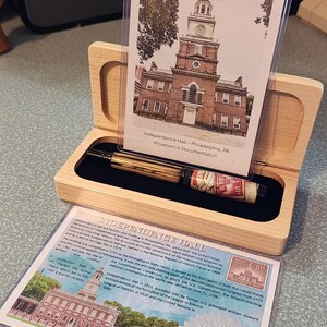 May include: A wooden pen box with a pen and documentation about Independence Hall in Philadelphia. The pen has a wood grain barrel and a red and white label. The documentation includes a photo of Independence Hall.