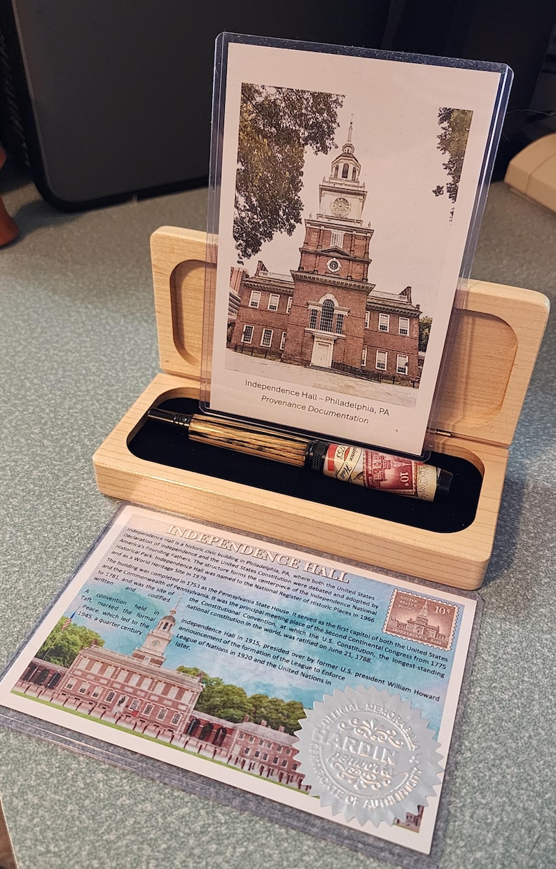 May include: A wooden pen box with a pen and documentation about Independence Hall in Philadelphia. The pen has a wood grain barrel and a red and white label. The documentation includes a photo of Independence Hall.