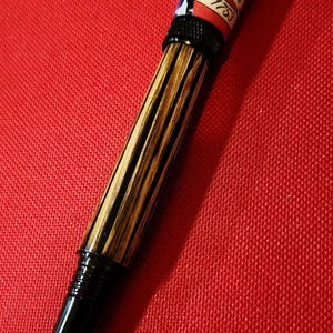 May include: A fountain pen with a black nib and a barrel featuring a wood grain design. The pen's upper barrel displays a vintage United States postage stamp design with the text "Independence 1753".