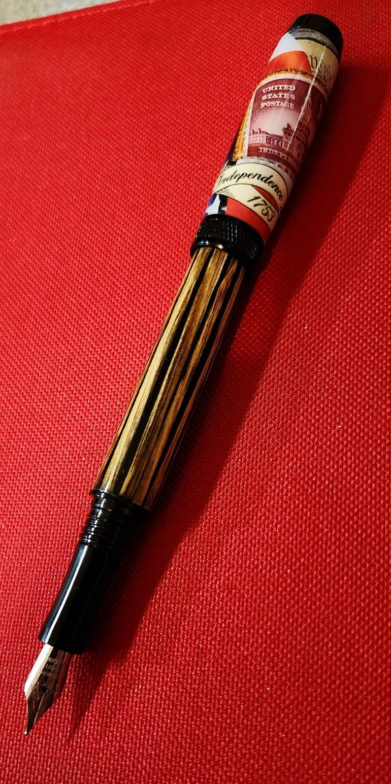 May include: A fountain pen with a black nib and a barrel featuring a wood grain design. The pen's upper barrel displays a vintage United States postage stamp design with the text "Independence 1753".