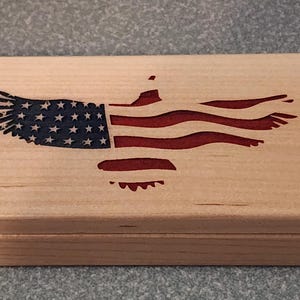 May include: A rectangular wooden box with a lid, featuring an engraved American flag design. The flag is depicted as an eagle with the stars and stripes in red, white, and blue. The box is made of light-colored wood.
