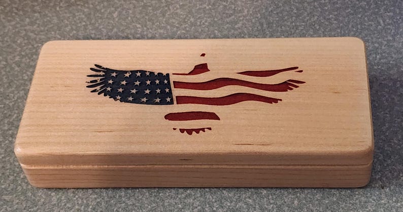 May include: A rectangular wooden box with a lid, featuring an engraved American flag design. The flag is depicted as an eagle with the stars and stripes in red, white, and blue. The box is made of light-colored wood.