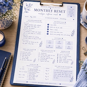 Peut inclure: Un presse-papiers bleu avec un planificateur de réinitialisation mensuelle imprimé, avec des sections pour la réflexion, les objectifs et les habitudes. Le texte comprend "Monthly Reset" et "Progress over perfection." Un cahier bleu, un stylo et une bougie sont également visibles.