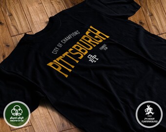 May include: Black t-shirt with a gold graphic that says "Pittsburgh" and "City of Champions" with a small white graphic of a baseball bat and ball. The shirt is made of super soft ringspun cotton.