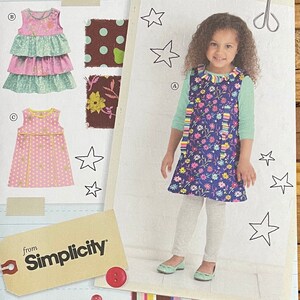 May include: A young girl wearing a blue floral print dress with a rainbow striped collar and straps. The image also shows a pink polka dot dress, a tiered dress, and fabric swatches. The Simplicity logo is visible.