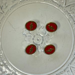 May include: Four small, oval red and gold buttons. Each button features a gold lightning bolt design. The buttons are arranged in a square pattern on a white, patterned surface.