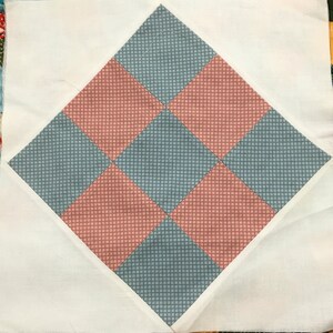 May include: A quilt block with a diamond pattern in shades of blue and pink. The block is made of fabric with a small, repeating pattern.
