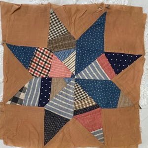 May include: A handmade quilt block featuring a star design. The star is composed of various patterned fabrics, including plaid, stripes, and polka dots, in shades of blue, red, and brown. The star is set against a solid brown background.