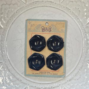May include: A set of four dark navy blue buttons with a geometric, faceted design and two center holes, mounted on a vintage cardboard display card labeled B.W.C. and Muscatine, Iowa, The World's Biggest Button Market, with handwritten text.