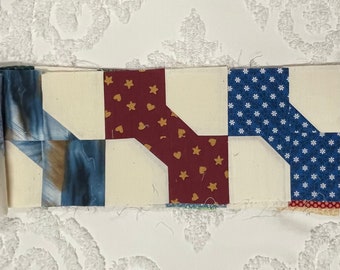 May include: A quilt block with a patchwork design featuring red, white, and blue fabric. The block is made up of four squares, each with a different pattern. The fabric is a mix of solid colors and prints.