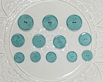 May include: A selection of teal buttons in different sizes, displayed on a clear, decorative plate. The buttons are glossy and have two holes for sewing. The plate features a floral pattern around its edge.