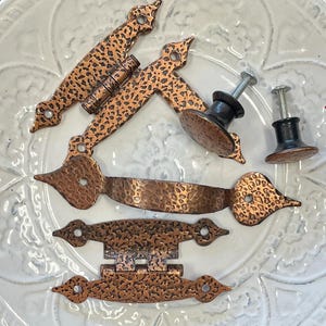 May include: A collection of copper-colored cabinet hardware, including handles, knobs, and hinges. The hardware has a hammered texture and a dark finish. The pieces are arranged on a white, patterned surface.