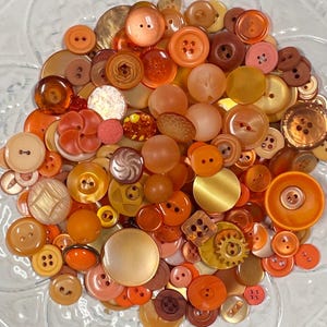 May include: A large collection of assorted buttons in various shades of orange, yellow, and brown. The buttons vary in size, shape, and texture, including round, square, and gear-shaped designs with two or four holes, featuring smooth, matte, and glossy finishes.