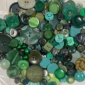 May include: A selection of green buttons in diverse shapes and sizes. The collection features a variety of shades, from light to dark green, including round, square, and textured designs, creating a visually interesting assortment.