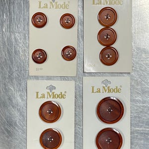May include: Four cards with brown buttons on them. Each card has the text "La Mode" and a price. The buttons are different sizes and have a wood grain pattern.
