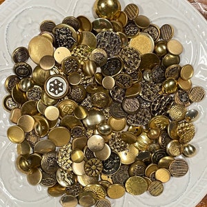 May include: A large collection of gold-toned buttons of various sizes and designs. The buttons are arranged in a pile on a white surface.