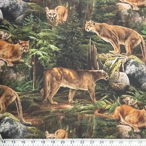 May include: A fabric print depicting several cougars in a forest environment. The cougars are shown in different sizes and positions, set against a backdrop of green trees, rocks, and foliage. The print has a realistic style with a ruler along the bottom edge.
