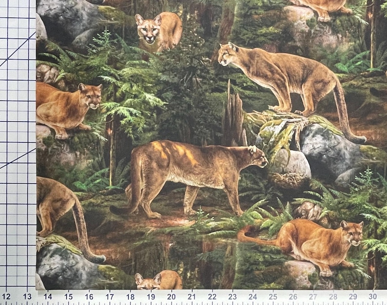 May include: A fabric print depicting several cougars in a forest environment. The cougars are shown in different sizes and positions, set against a backdrop of green trees, rocks, and foliage. The print has a realistic style with a ruler along the bottom edge.