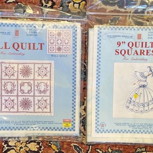 May include: Two vintage embroidery kits. One is a wall quilt pattern with geometric designs, and the other is a 9-inch quilt square featuring a parasol lady design. Both kits are in plastic packaging with blue and white borders.