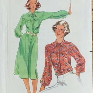 May include: Two women wearing blouses with bow ties. The woman on the left is wearing a green dress with a button-up front and a belt. The woman on the right is wearing a red and white floral blouse and a white skirt.