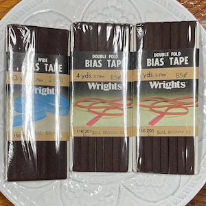 May include: Three packages of brown bias tape are displayed. The packages have text that reads "WIDE S TAPE" and "DOUBLE FOLD BIAS TAPE". The packages are labeled "Wrights" and include measurements in yards.