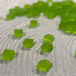 May include: A collection of small, translucent, lime green square tiles with a smooth, glossy finish. The tiles are scattered across the frame, with some grouped together in a cluster and others dispersed individually across the textured surface.