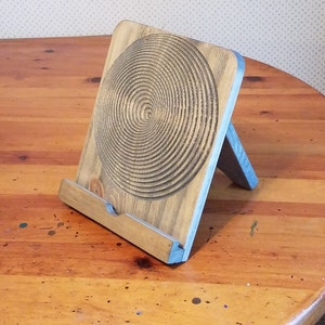 May include: Wooden tablet stand with a spiral design carved into the front. The stand is made of light-colored wood with a blue accent on the edges. The stand is angled and has a small ledge to hold a tablet or book.