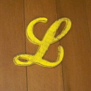 May include: A yellow wooden letter L, painted in a distressed style, is mounted on a wood panel background.