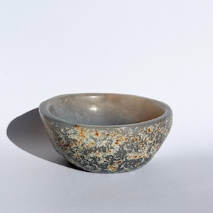 May include: A small, rounded bowl carved from translucent grey stone. The exterior features a natural, speckled pattern with clusters of white and rusty orange inclusions embedded within the smooth, polished surface of the mineral material.