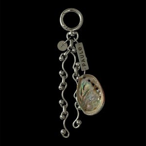 May include: A silver-toned keyring featuring a circular ring, a small round charm with the letter T, and a rectangular tag engraved with the name PAULE. Two wavy silver chains with spherical accents hang alongside an iridescent abalone shell.