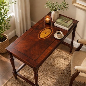 May include: A rectangular, dark wood accent table with an oval inlay design. The table has four ornate legs and is topped with a vase of greenery, books, a candle, and a cup of coffee. The table is 48 inches long.