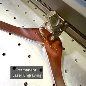 May include: Close-up of a laser engraving process on a brown leather object. The text "Permanent Laser Engraving" is displayed. The laser etches the surface, creating a detailed design. The object is on a metal surface.