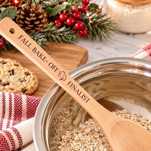 May include: A light-colored wooden spoon with a hole in the handle, engraved with the text "FALL BAKE-OFF FINALIST" and a small chef hat icon. The spoon is positioned inside a stainless steel mixing bowl containing dry ingredients.